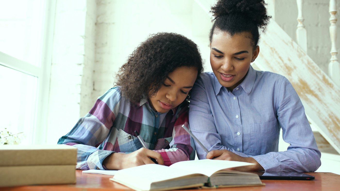 A mother and her teenage daughter working through a book together at a table